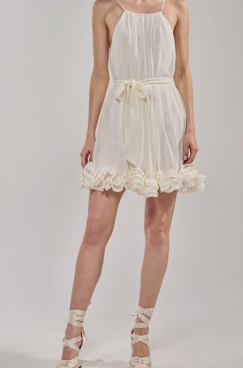 Video of woman modeling the CAMERA PLEASE RUFFLE MINI DRESS—an ivory, sleeveless mini dress with spaghetti straps, pleated texture, waist tie, and ruffled hem—posing against a light background. Perfect for summer occasions.