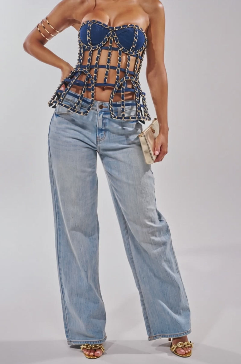 A video of a woman in the DOWN SOUTH CHAIN DETAIL BUSTIER TOP, paired with wide-leg light blue jeans, gold sandals, and a beige clutch against a plain light background.