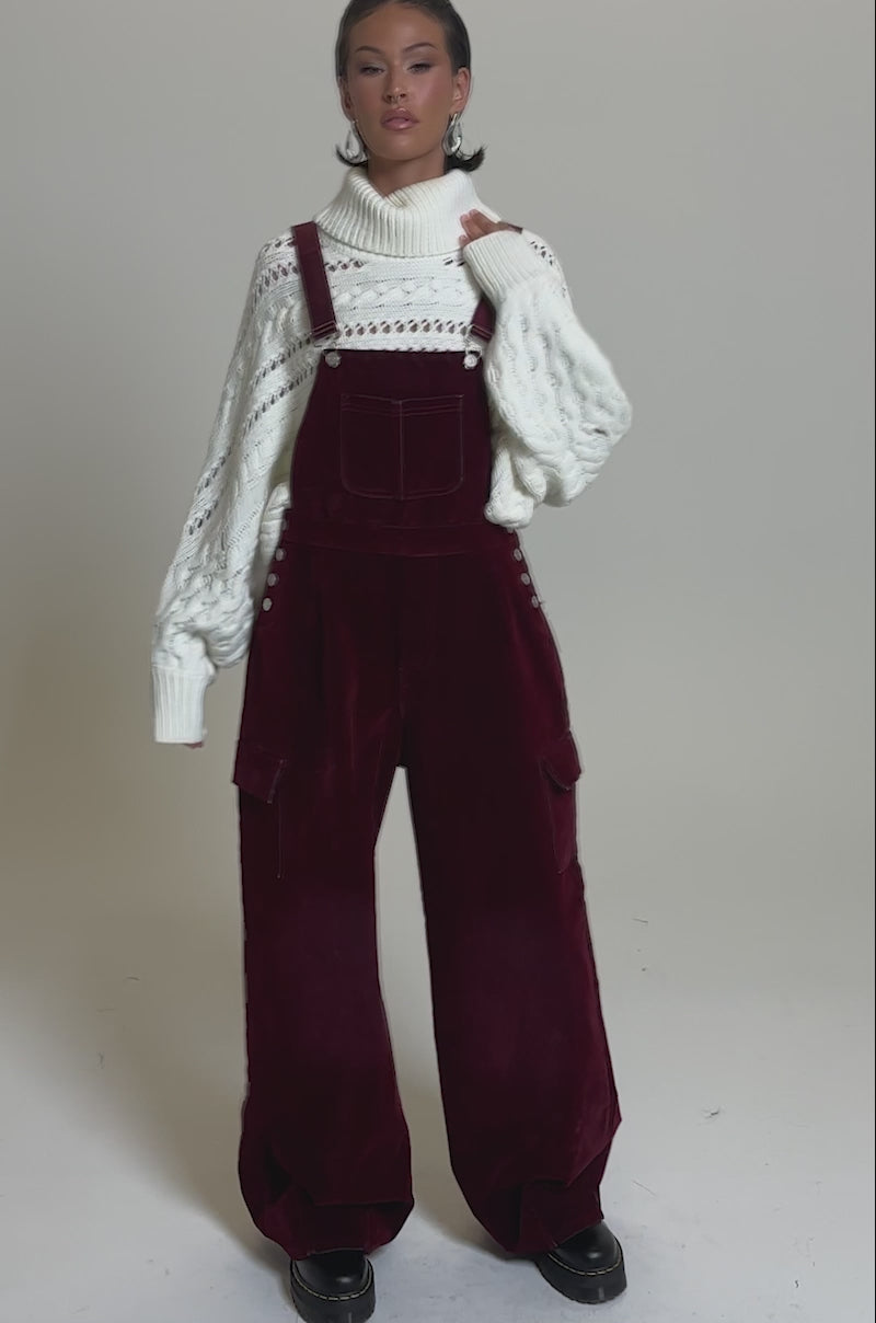 A video of a model wearing the IT'S GIVING VINTAGE VELVET DENIM OVERALLS with a wide-leg silhouette and a chunky white turtleneck stands confidently against a plain gray background.