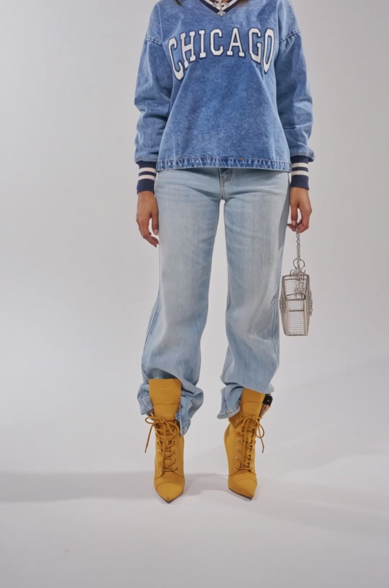 A video of a woman poses in light blue jeans, a CHICAGO SWEATSHIRT, mustard yellow boots, sunglasses, a striped cap, and holds a small silver handbag.