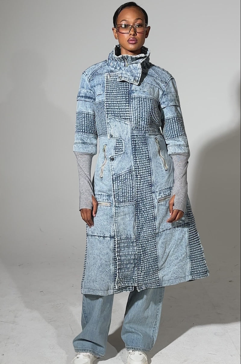 A video of a person in the VORTEX DENIM TRENCH COAT IN BLUE, sunglasses, light blue jeans, and white sneakers stands against a plain light background.