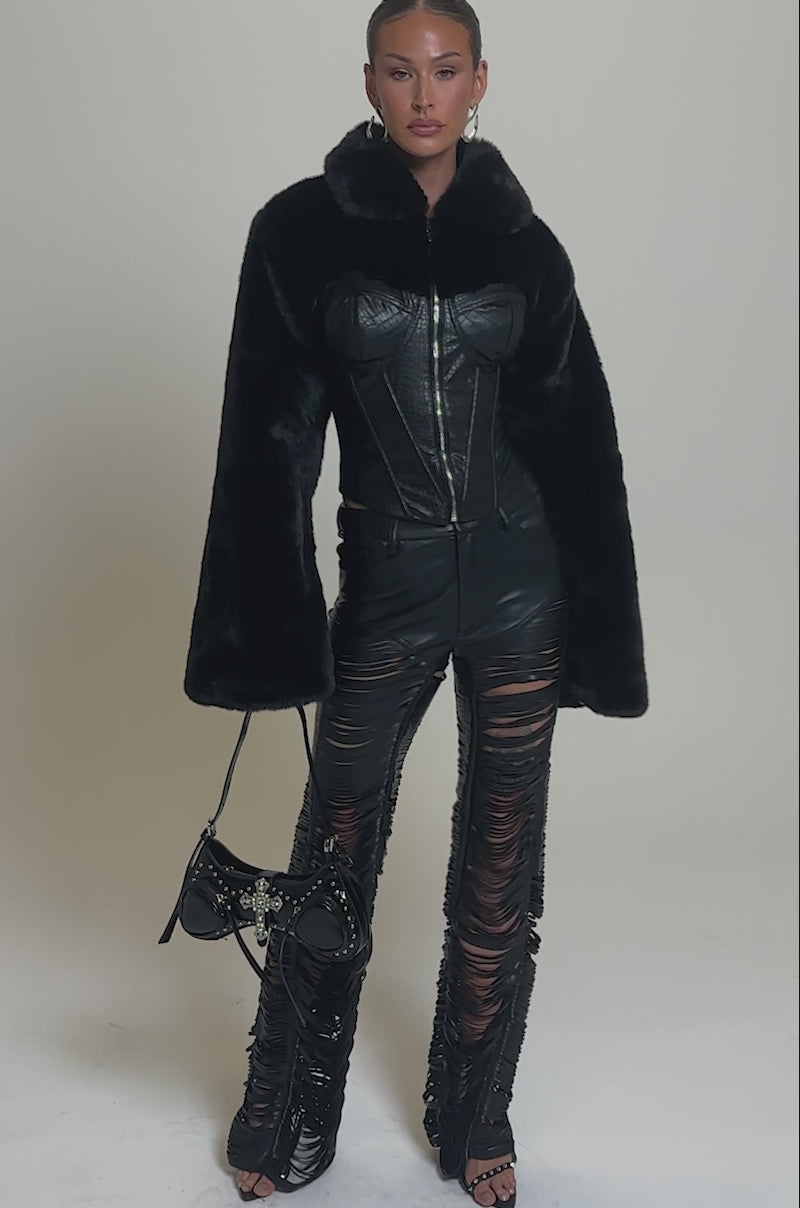 Video of woman modeling the SHORTY CORSETED FAUX FUR jacket, featuring a cropped fit and corset bodice, paired with ripped leather pants and a black handbag against a neutral background.