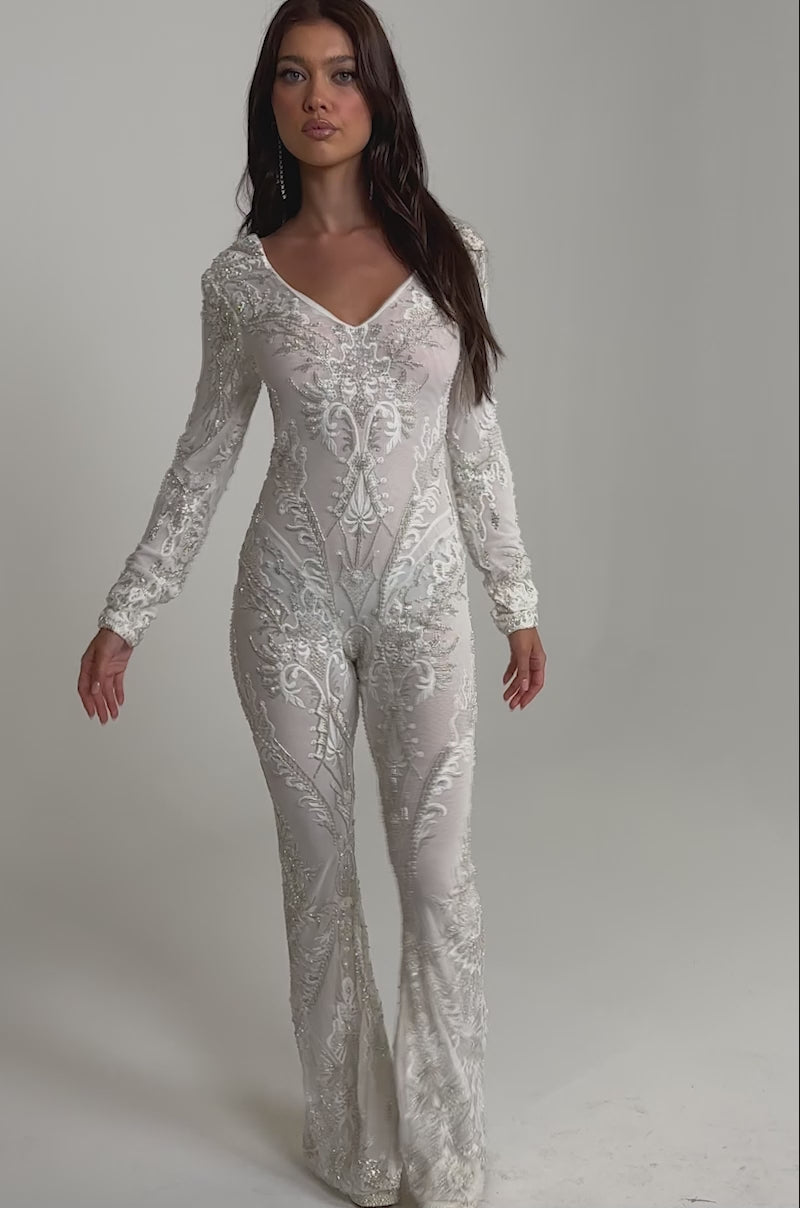 Video of woman modeling the WATCH ME WORK EMBELLISHED SEQUIN JUMPSUIT IN WHITE, featuring a fitted silhouette, flared legs, and sparkling sequin details. She has long brown hair and neutral makeup against a plain light background.