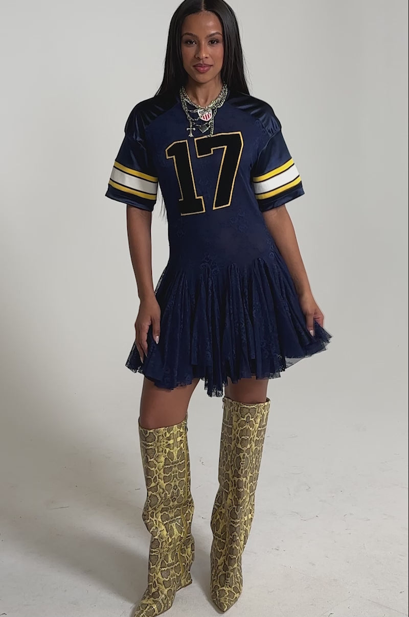 A video of a woman in the VARSITY BLUES LACE MINI DRESS with tall yellow snakeskin pointed-toe boots.