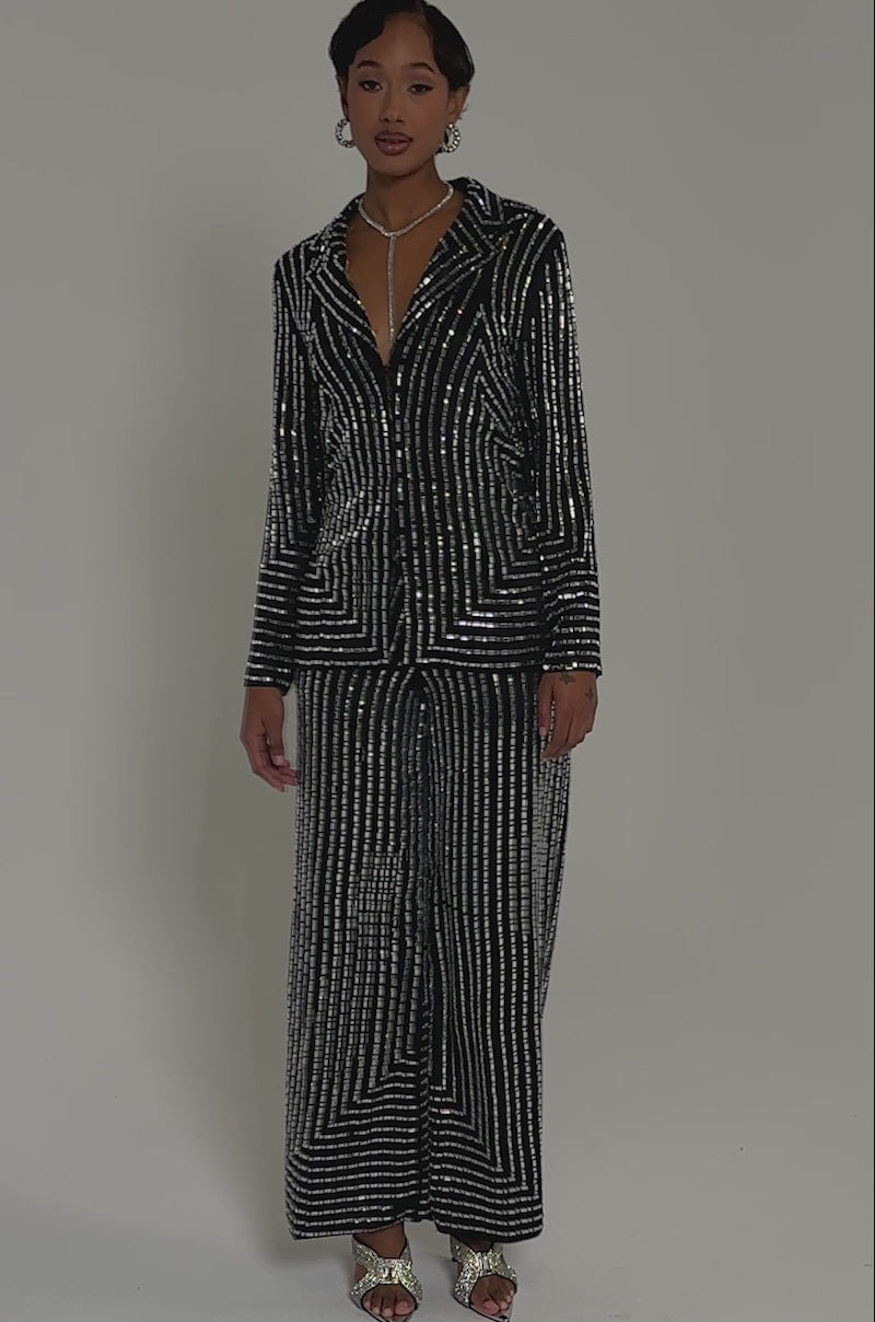 Video of woman modeling the DONT STARE TOO HARD RHINESTONE TROUSER, featuring black sequins, geometric patterns, and a high-rise wide-leg fit, paired with silver heels and a statement necklace against a light gray background.