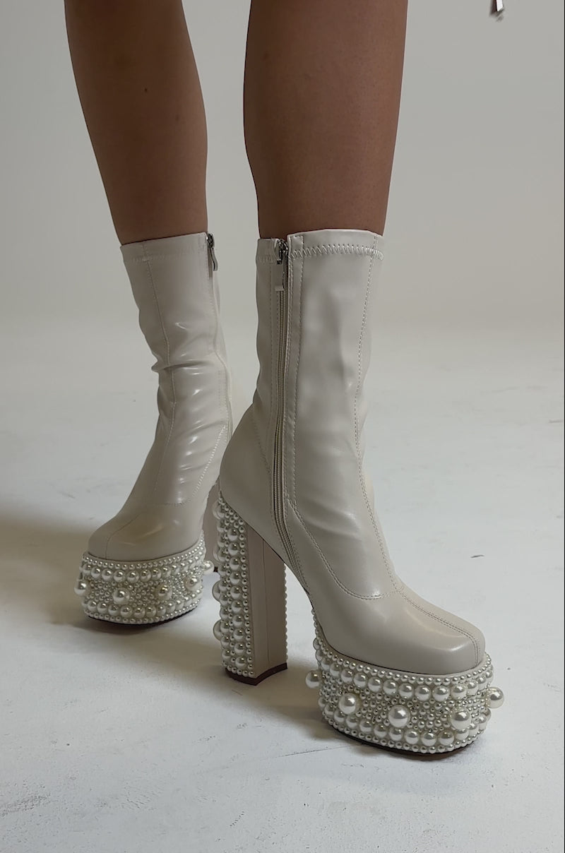 A video of the AZALEA WANG ANTARIS BEIGE PEARL DETAIL PLATFORM BOOTIE featuring cream faux leather, chunky heels, and platform soles embellished with rows of white pearls, as shown worn on a light-colored surface.