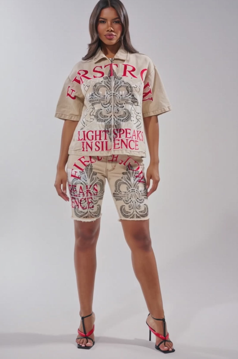 A video of a woman in the LIGHT SPEAKS IN SILENCE DENIM SHORT, beige with bold black patterns and red text, paired with a matching zip-up shirt and black and red strappy heels.