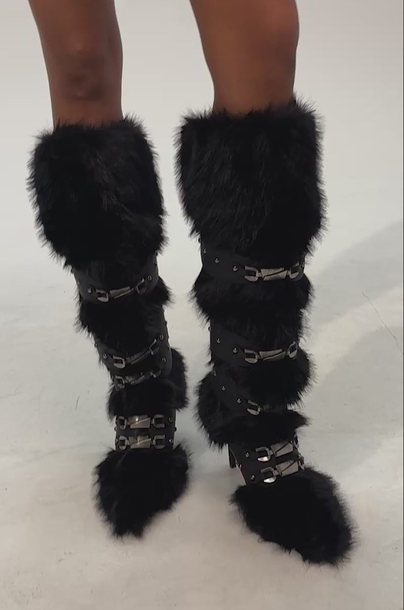 Video of woman modeling the AKIRA GOLD DARIO BLACK FUR WITH STRAPS KNEE HIGH BOOT—sexy black stiletto boots featuring faux fur, multiple straps, and metallic details—against a plain light background.