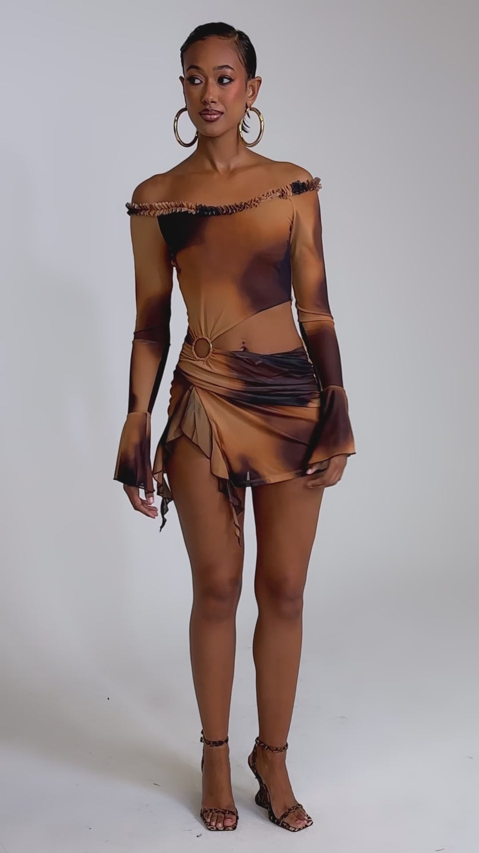 A video of a woman in the GIVE IT TO ME BABY MINI DRESS, a fitted off-shoulder mesh mini with brown and orange ombre, side cutout, ruffled hem—styled with hoop earrings and heels—perfect for making a statement at any party.