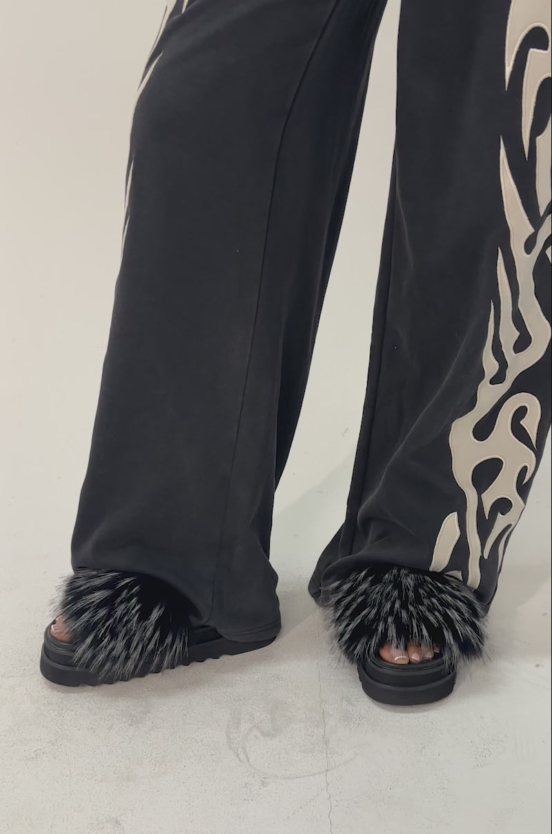 A video of a person wearing black pants with white abstract patterns and the AKIRA MIRAY BLACK MULTI BLACK FUR SLIDE sandals.