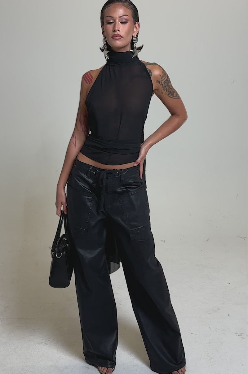 A video of a woman in black wide-leg pants and the GOT IT FROM YOU BLOUSE—a black halter wrap top with draped fabric—paired with large star earrings, a stylish black handbag, and wet-look hair against a plain light background.