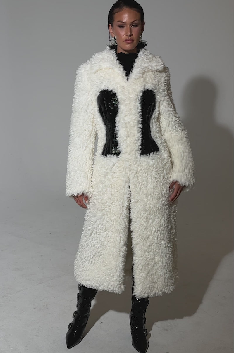A video of a woman posing in the BUY ME ROSES CORSETED FUR COAT, a long white fur style with black corset detailing, paired with black textured boots. Her hair is slicked back as she stands against a plain light background.