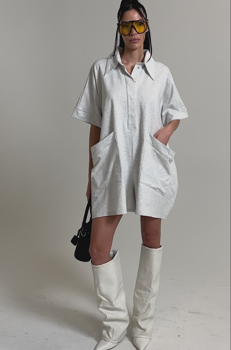 A video of a woman in the SHE'S RICH OVERSIZED FRENCH TERRY ROMPER with large pockets, styled with white knee-high textured boots, yellow-tinted sunglasses, and a gray clutch against a plain background.