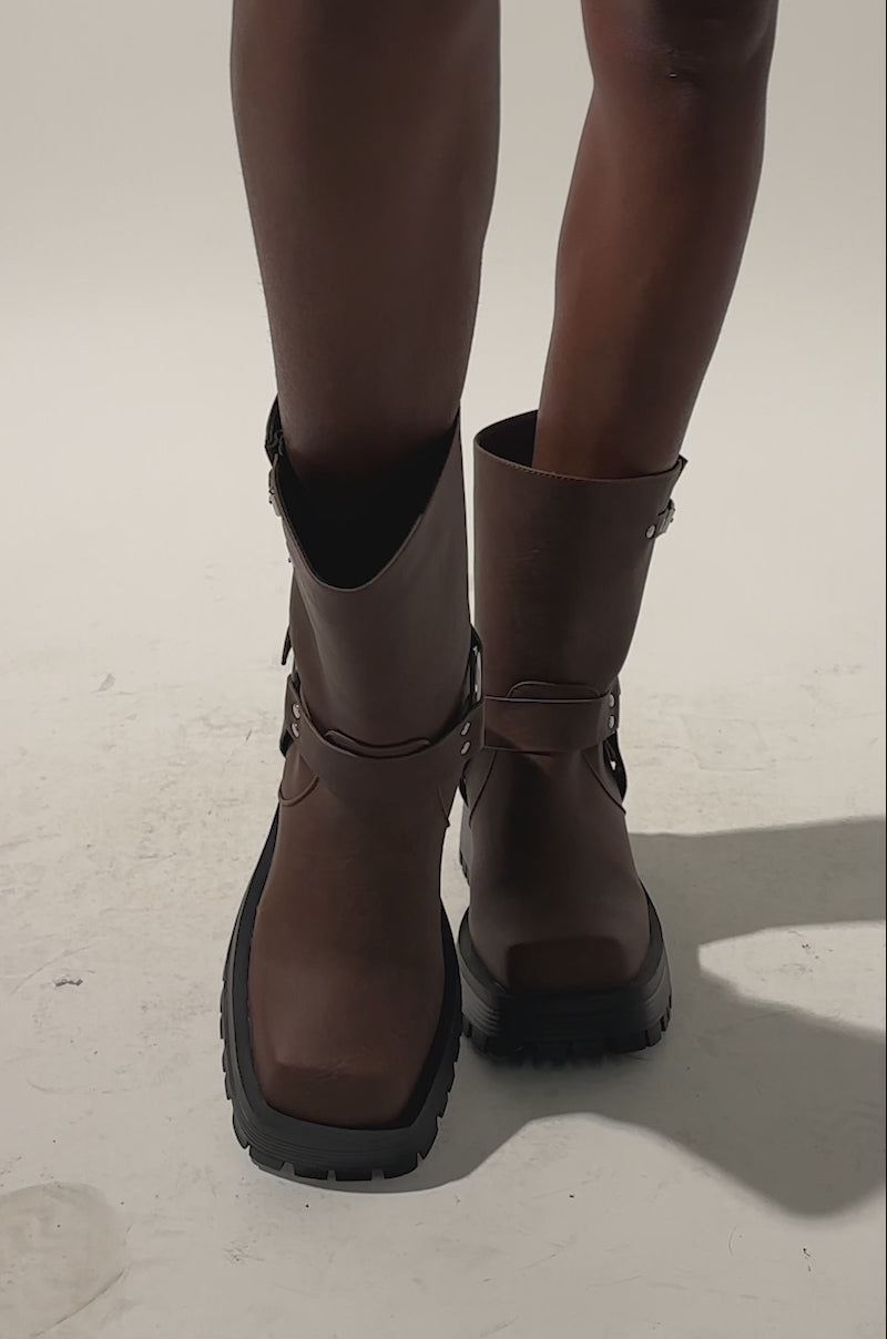 A video of a person with medium-brown skin wearing the AZALEA WANG BUCKED BROWN CHUNKY HEEL COMBAT BOOTIE, featuring brown faux leather, black chunky soles, and silver buckles, standing on a light background.