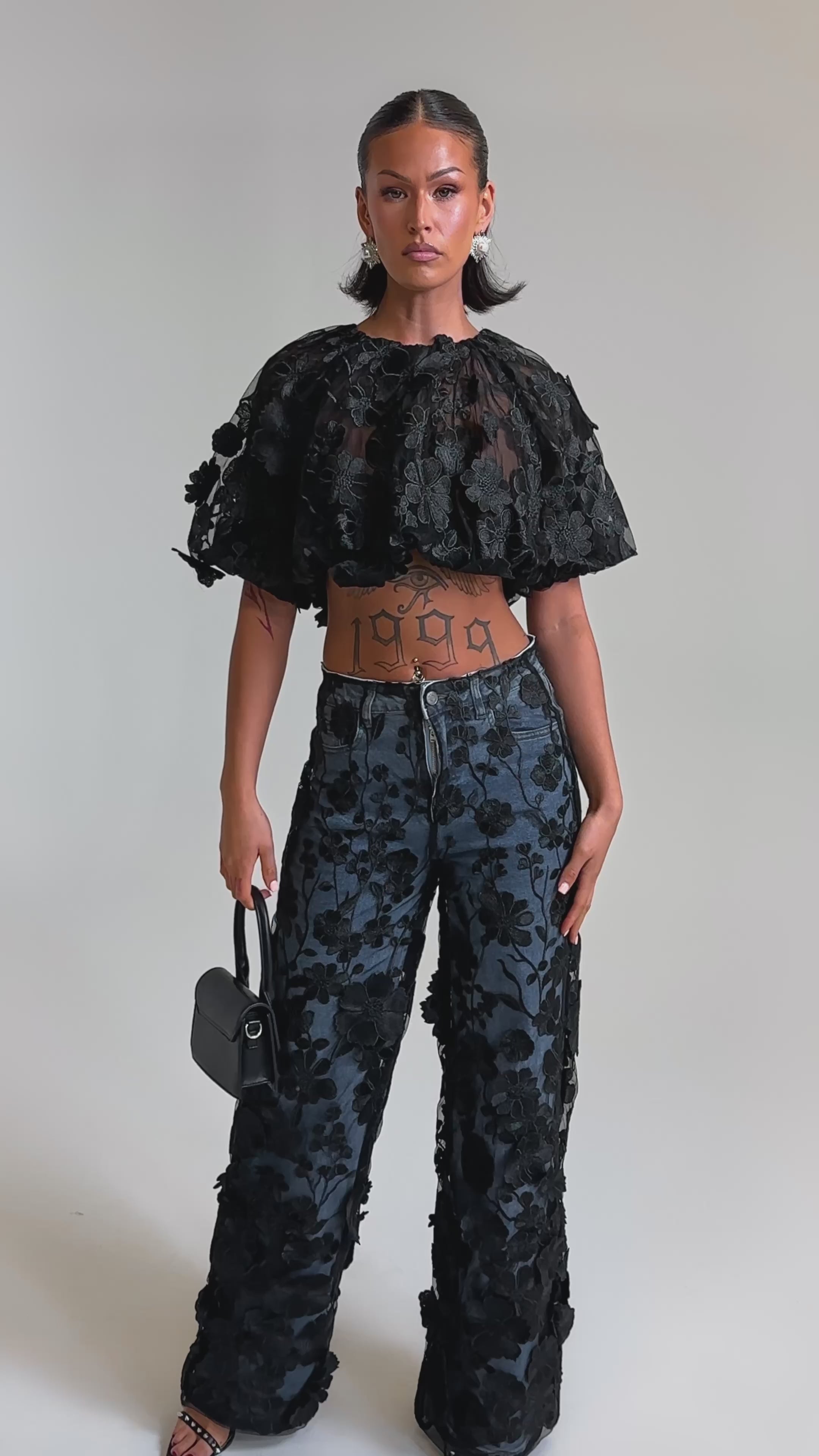 A video of a woman in a black textured cropped top, open-toed heels, and the DONT GIVE UP RELAXED DENIM WITH TULLE OVERLAY. She carries a small black handbag and wears her hair slicked back.