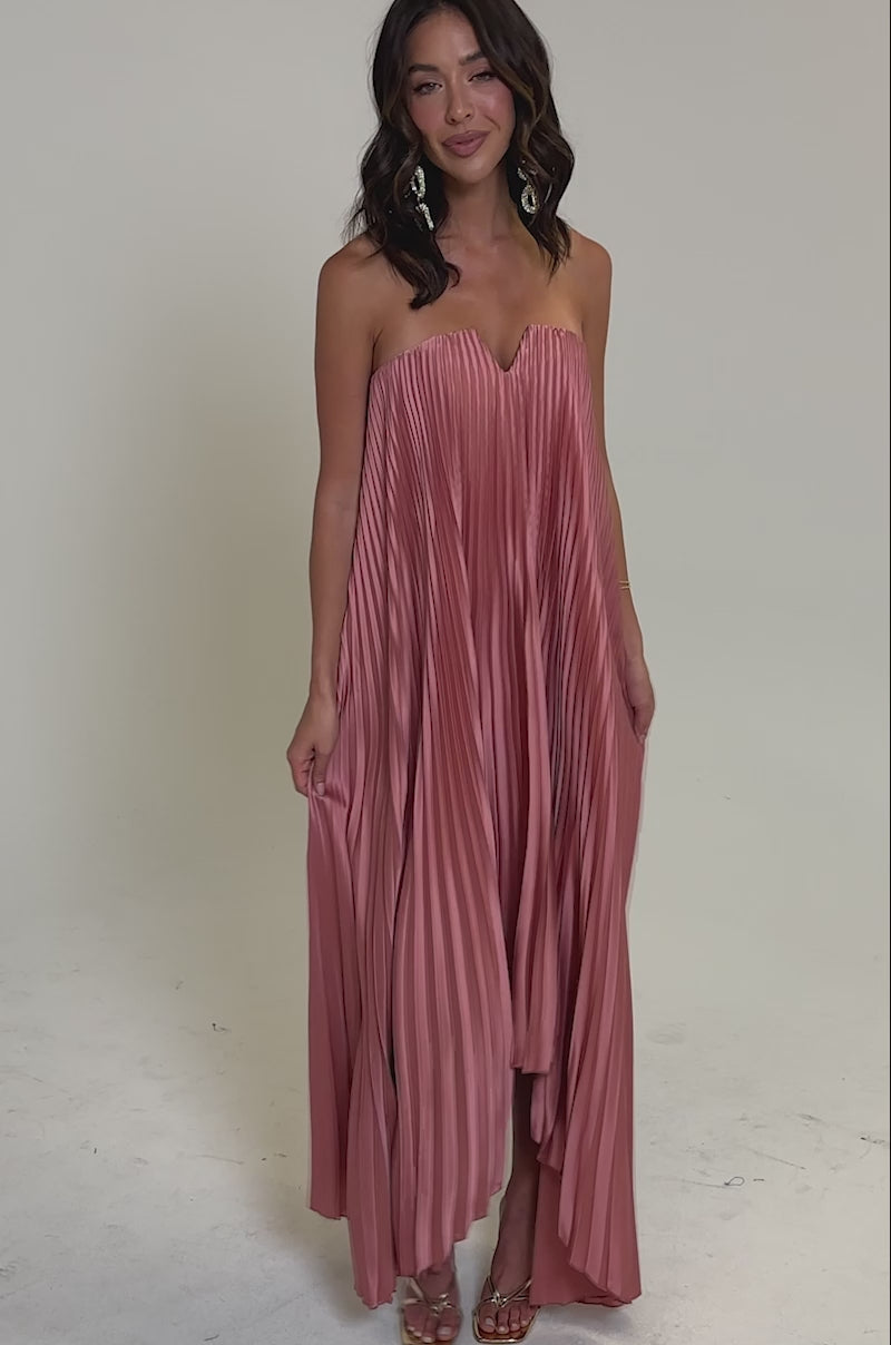 Video of woman modeling the EMERALD DREAM SLEEVELESS MAXI DRESS IN PINK, a pleated flowy pink formal maxi. She wears gold sandals, hoop earrings, a bracelet, and gently holds the skirt with her wavy brown hair loose.