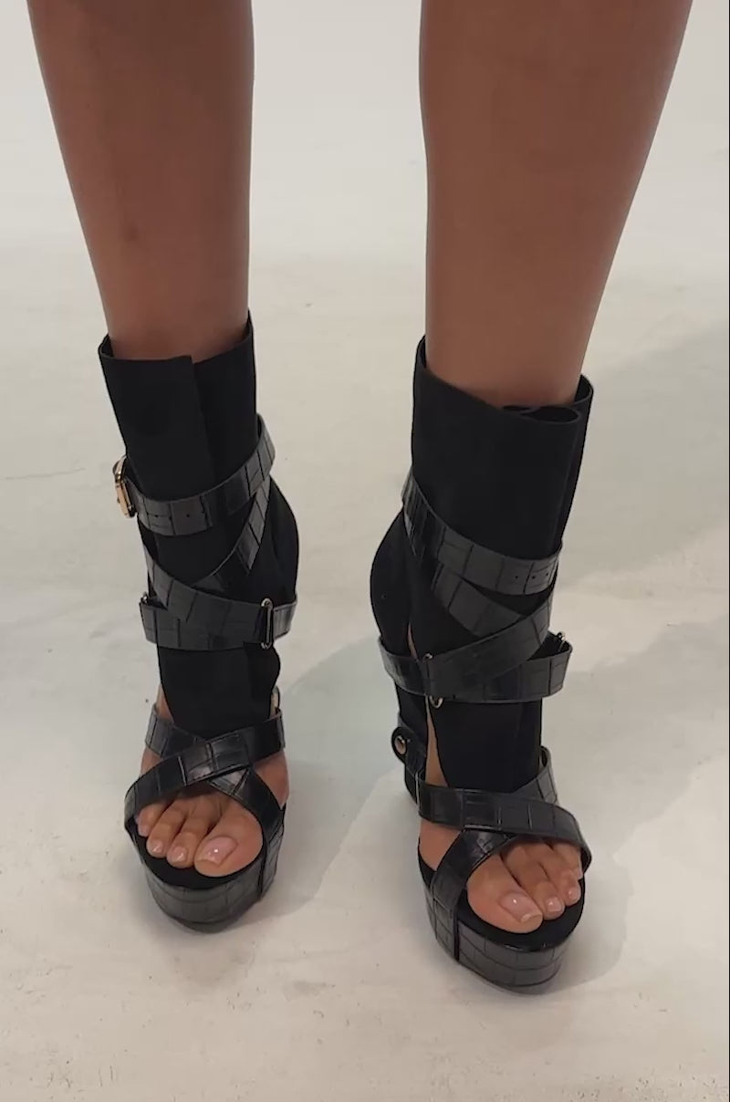 Video of woman modeling the AZALEA WANG ROTHKO WEDGE SANDAL IN BLACK featuring a high ankle cuff, multiple crisscross straps, a gold buckle, and a textured platform sole