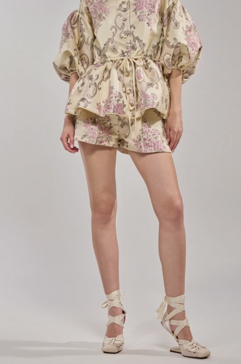 Video of woman modeling the PORCELAIN DOLL BROCADE SHORTS: high-rise cream shorts with pink and taupe floral print, pleats, and side pockets