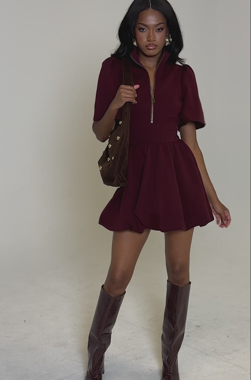 A video of a woman modeling the NAME OF THE GAME SHORT SLEEVE ZIPPERED MINI DRESS IN BURGUNDY, posed against a plain light gray background.