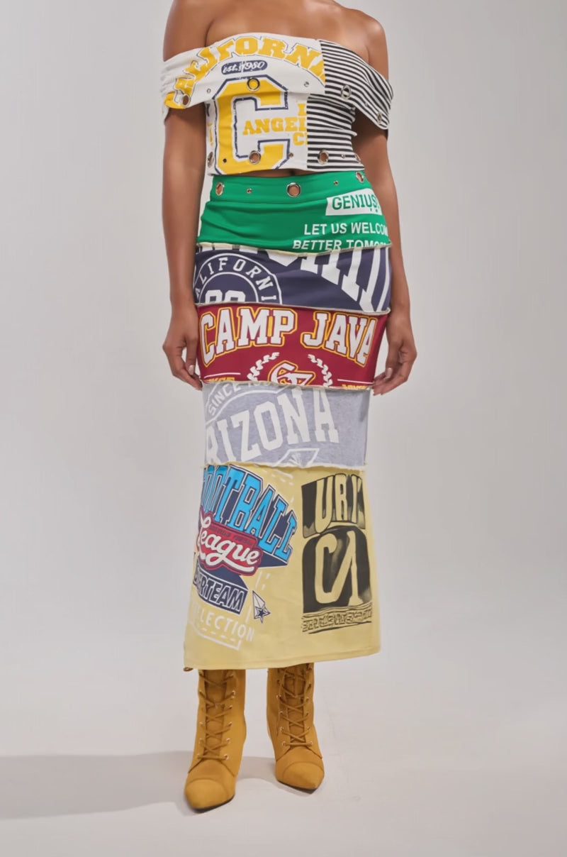 Video of woman modeling the FULL RIDE PATCHWORK MAXI SKIRT, crafted from colorful graphic T-shirts, paired with an off-shoulder top and tan lace-up boots. She has slicked-back hair and neutral makeup against a plain background.