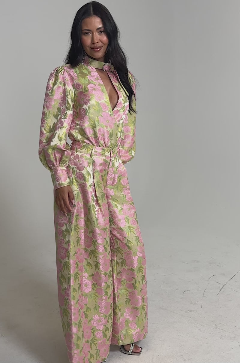 Video of woman modeling the OVER IT FLORAL BODYSUIT, a shiny long-sleeve one-piece with pink and green floral print, puffed sleeves, a deep V-neckline, and a choker neck with ring. She has long dark hair and smiles slightly.