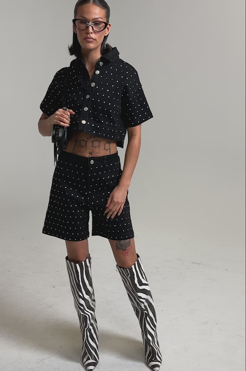 A video of a woman in the ITS GIVING BERMUDA SHORT, a black studded short-sleeve jacket, zebra-print knee-high boots, black purse, glasses, and hoop earrings.