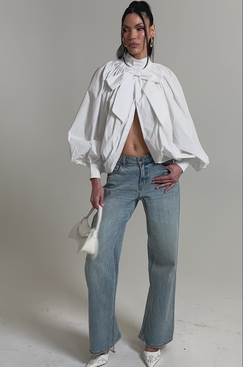 A video of a woman in the BLACK TIE AFFAIR BLOUSE IN WHITE with large bow details and puffy sleeves, paired with light blue wide-leg jeans, white pointed heels, and a small white textured handbag against a plain background.