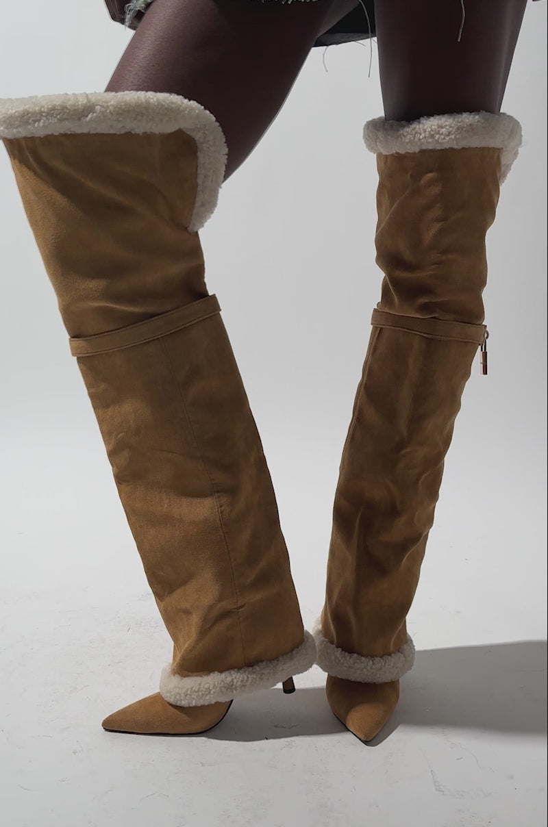 A video of a person wearing an asymmetrical denim mini skirt and oversized tan AZALEA WANG RACHANA TAN SHERPA STILETTO BOOTS with pointed toes and a small gold lock detail.