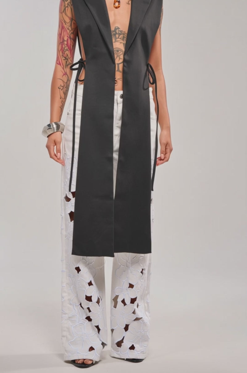 A video of a woman in the HONEY HONEY TIE VEST IN BLACK, an asymmetrical piece with deep lapels and a high-low hem, styled over white wide-leg floral cutout pants. 