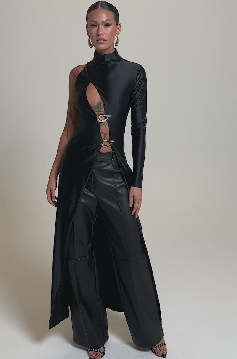 A video of a woman in a long black asymmetric top with metallic ring details, styled over FAUX STRETCHY LEATHER TROUSER. She has slicked-back hair, gold hoop earrings, and visible tattoos on one arm.