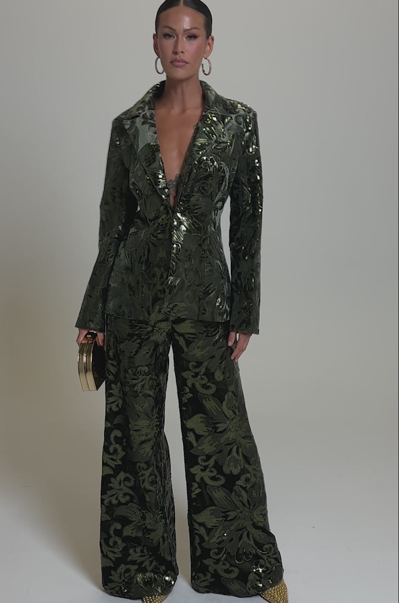 A video of  a person in a shiny patterned green pantsuit with wide-leg trousers and the AURORA SEQUIN VELVET BLAZER, accessorized with a gold clutch, gold pointed shoes, hoop earrings, and slicked-back hair.