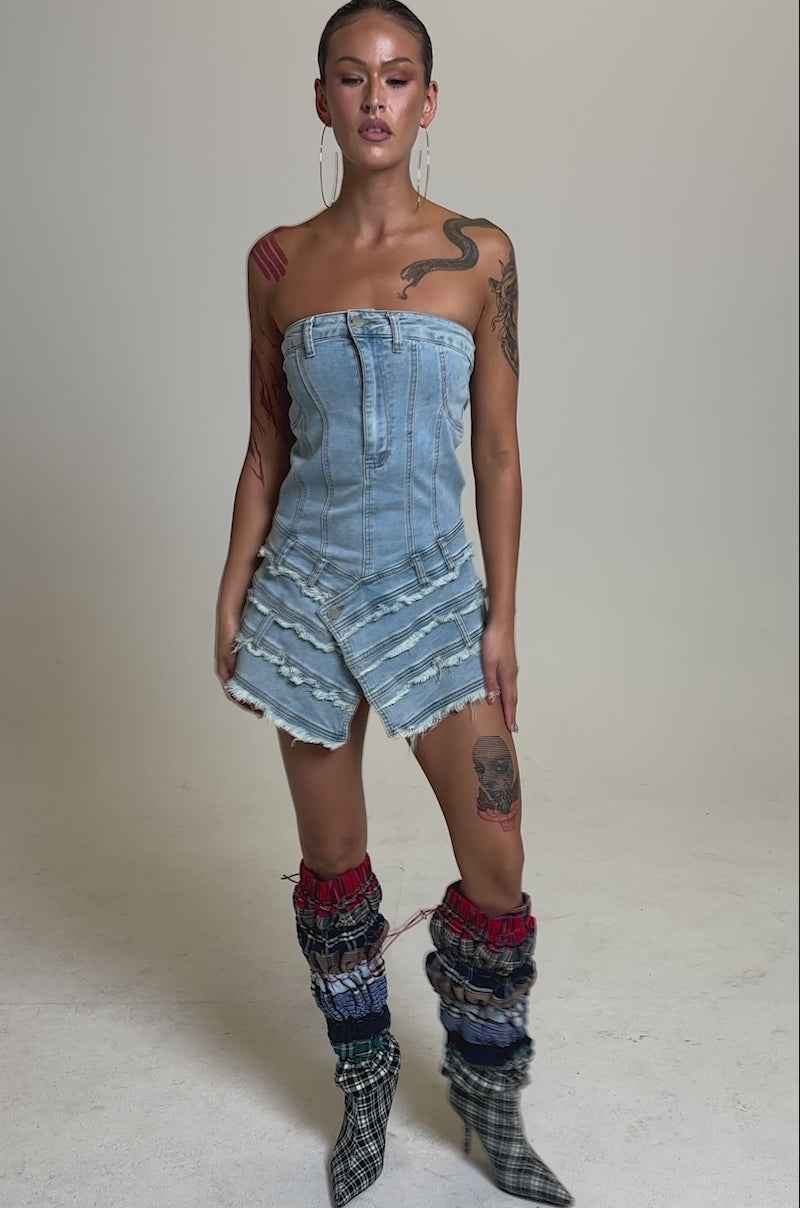 A video of a woman standing confidently in the strapless TOO MUCH TO LOVE DENIM ROMPER, paired with plaid heeled boots and colorful ruched leg warmers. Her hair is slicked back, and she has visible tattoos on her arms and leg.