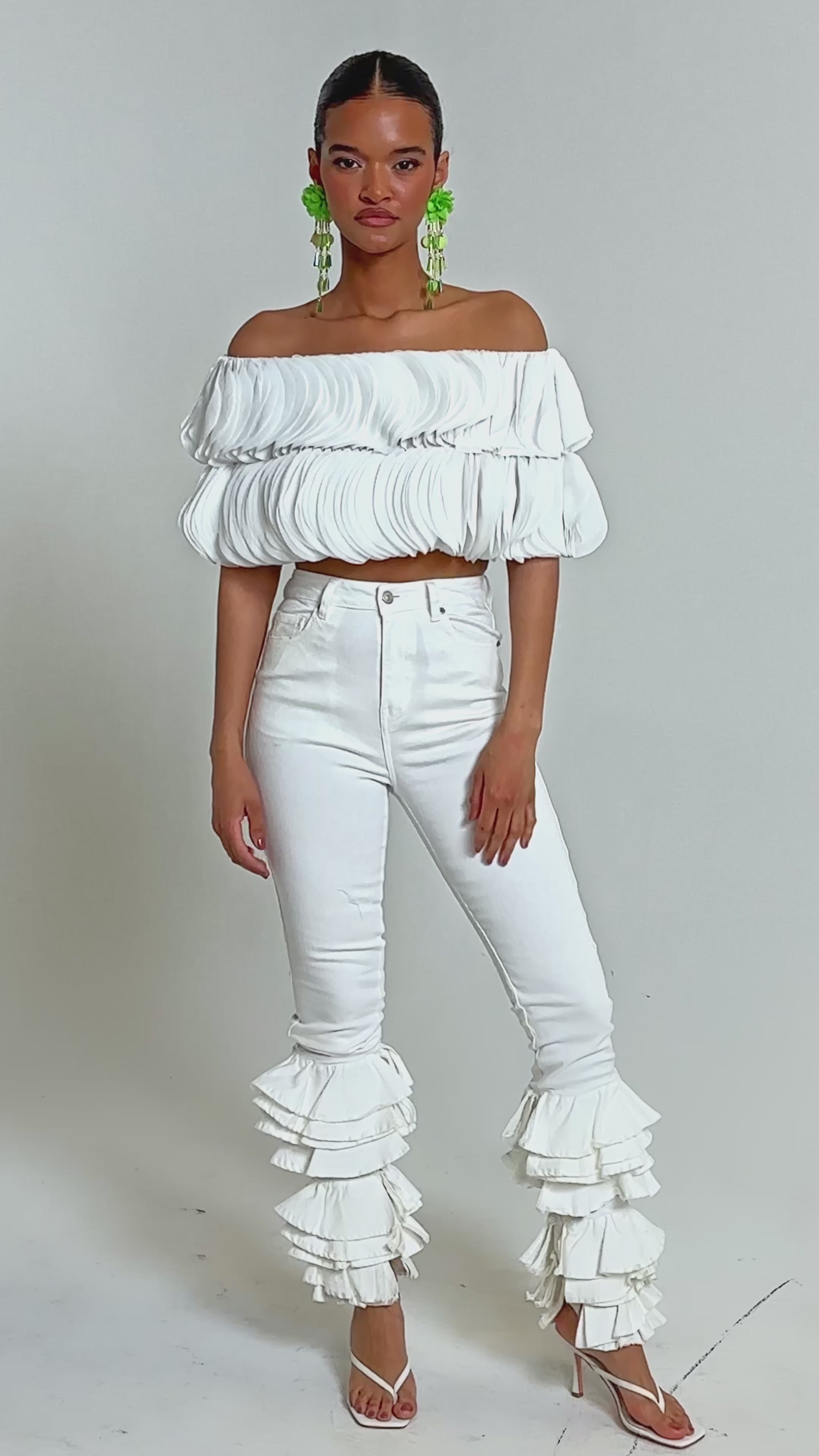 Video of woman modeling the DAYDREAMER CHIFFON BLOUSE, a white off-shoulder layered top, paired with high-waisted white pants. Her slicked-back hair and bold green earrings complete the editorial-inspired look against a light background.