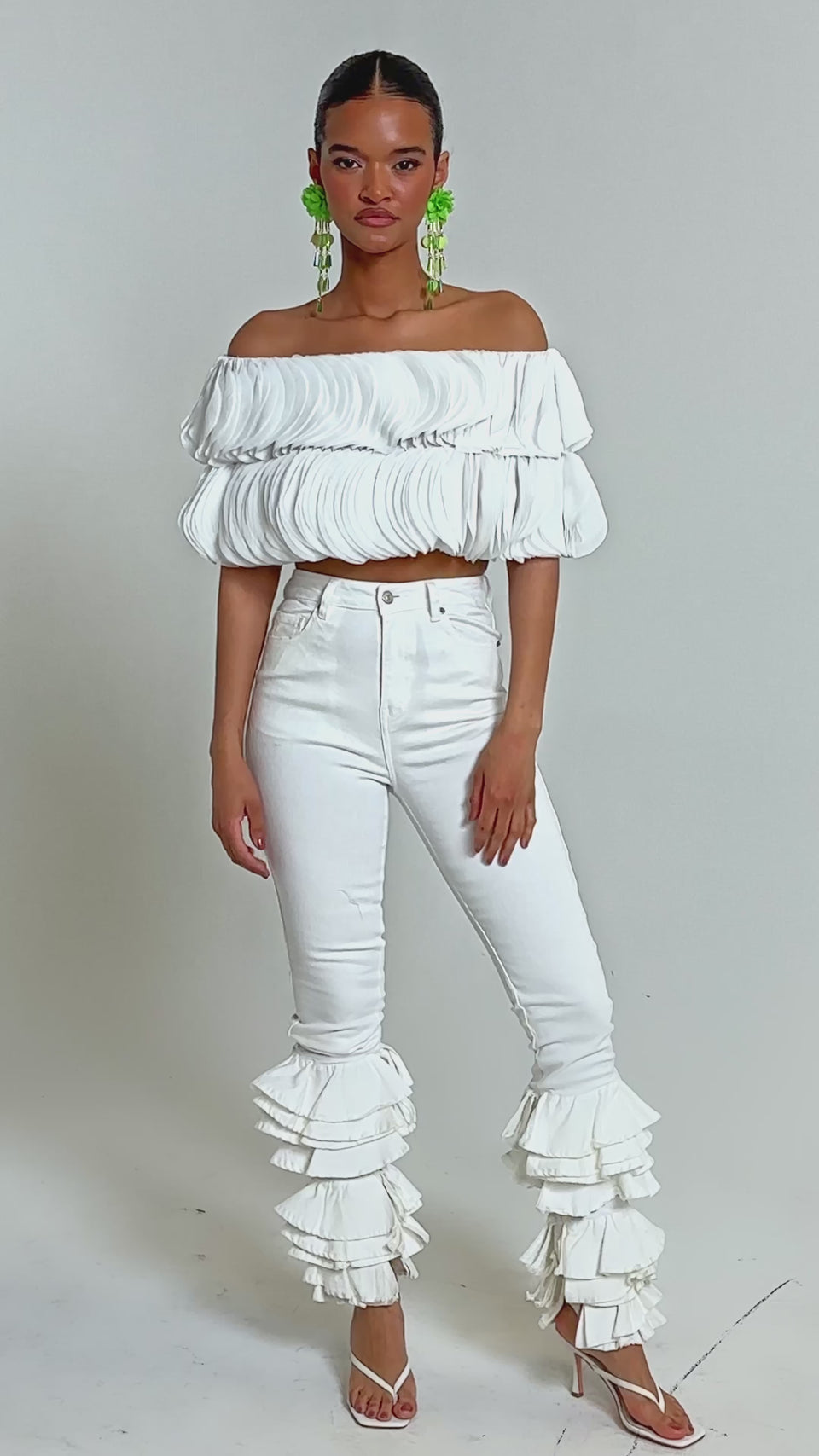 Video of woman modeling the DAYDREAMER CHIFFON BLOUSE, a white off-shoulder layered top, paired with high-waisted white pants. Her slicked-back hair and bold green earrings complete the editorial-inspired look against a light background.