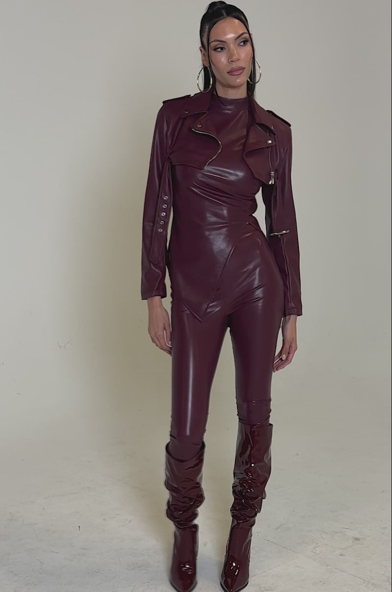 A video of a woman wearing the HARLEY LUXE ULTRA CROP MOTO IN BURGUNDY, a shiny faux leather jacket, paired with an asymmetric top, fitted pants, and knee-high boots against a plain light background.
