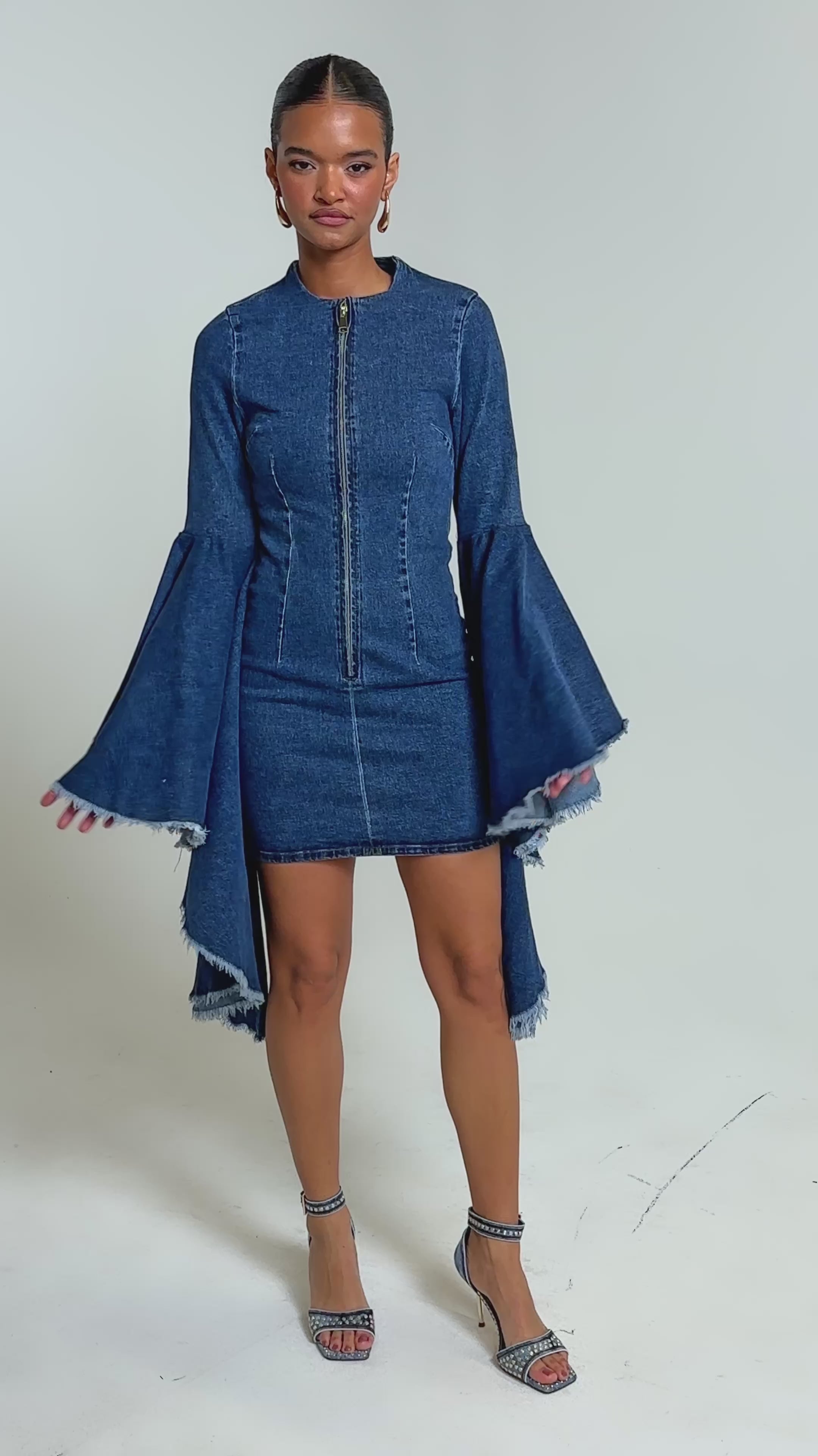 Video of woman modeling the WALK IN THE PARK DENIM MINI DRESS, featuring tiered bell sleeves and a frayed hemline. She pairs it with matching denim heels and styles her hair in a long braid.