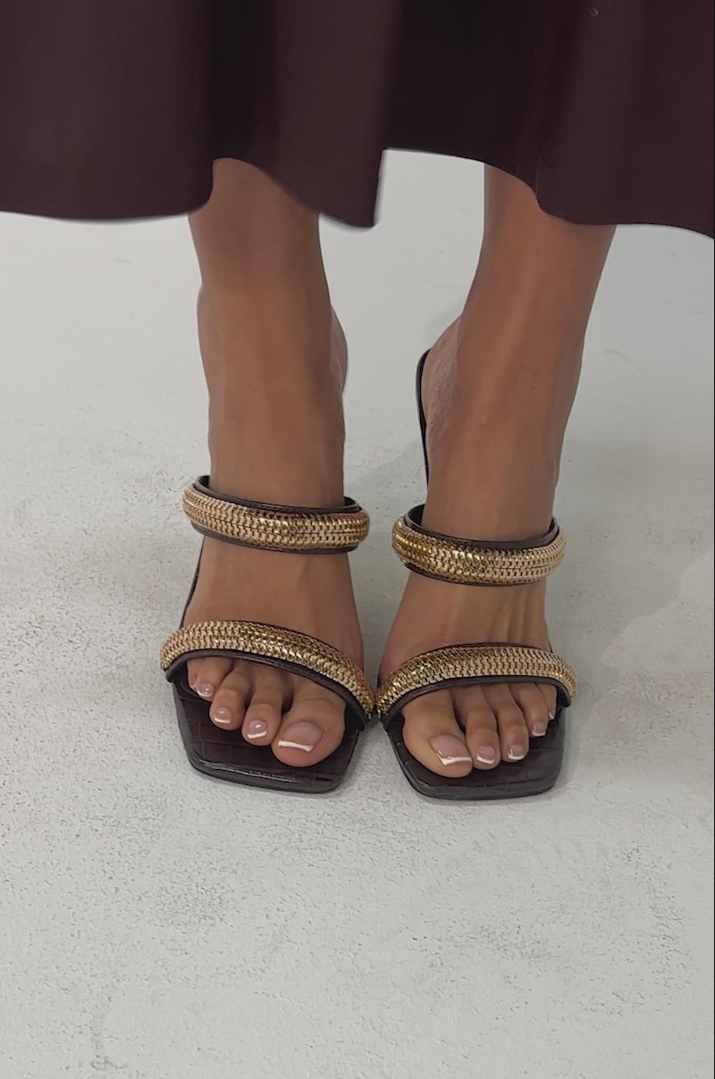Video of woman modeling the AZALEA WANG BESSIE CHOCOLATE CROC GOLD EMBELLISHED WEDGE SANDAL, featuring gold hardware straps and unique geometric gold heels, standing against a plain light background.