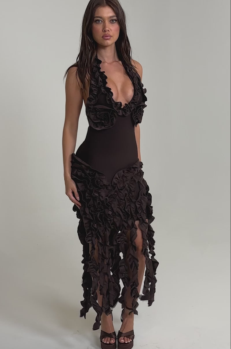 Video of woman modeling the STANDING OVATION FLOCKED MAXI DRESS IN CHOCOLATE—a sleeveless brown halter maxi with bold chest and skirt ruffles. She pairs it with brown heels, posing against a light plain background.