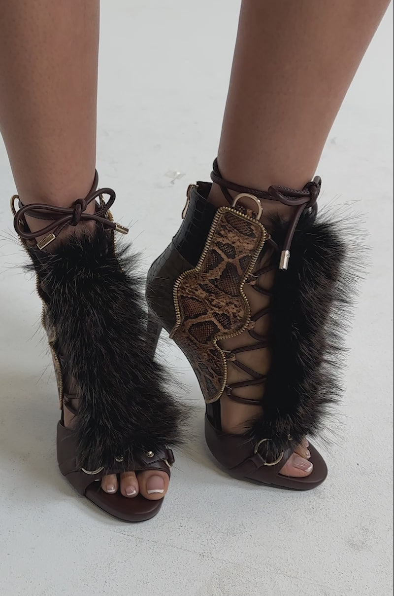 A video of a person wearing the AZALEA WANG SHAINY SNAKE BROWN MULTI sandals—open-toe, lace-up stiletto heels with brown faux leather, faux fur trim, and snakeskin accents.