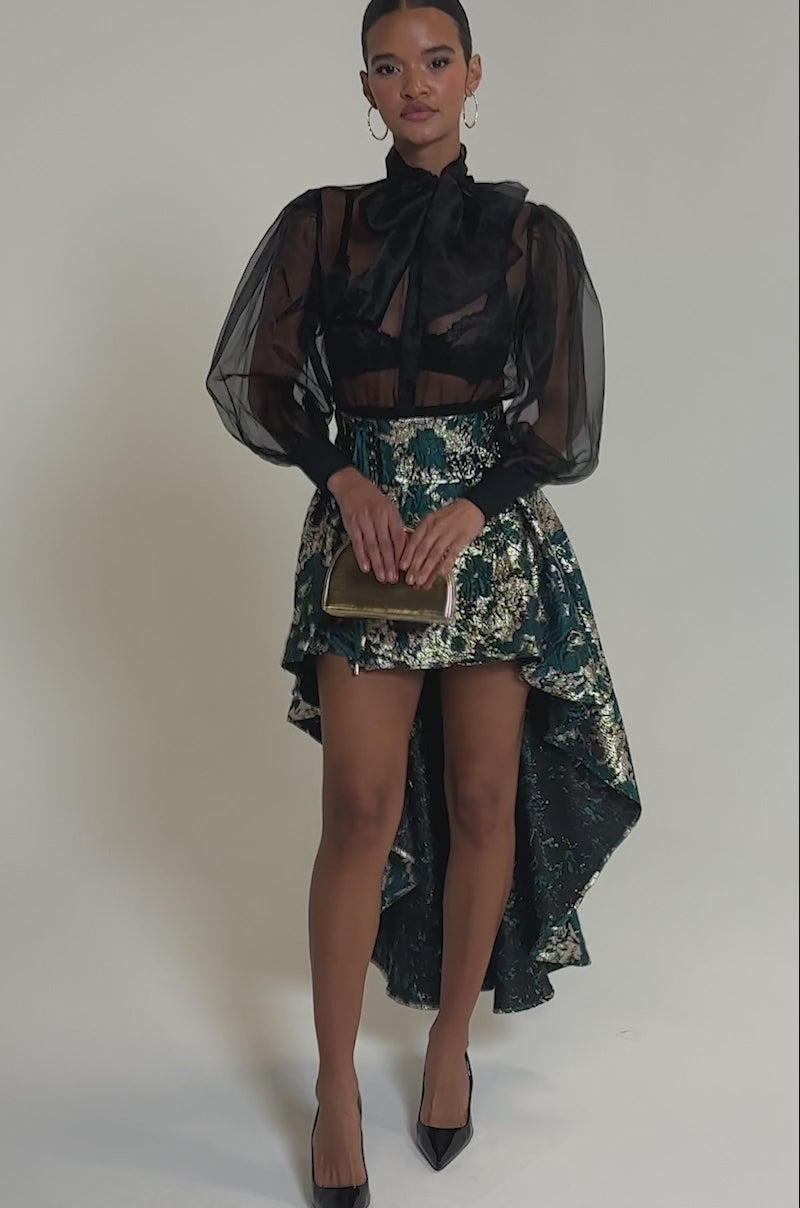 Video of woman modeling the GLORIOUS DAYS BROCADE HIGH LOW SKIRT in teal and gold with a sheer black blouse, large bow, black pointed heels, and a gold clutch.