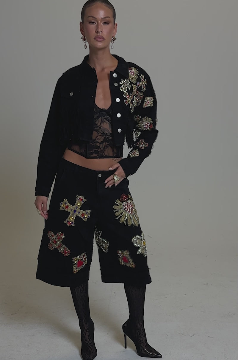 A video of a woman posing confidently in the MAIN SQUEEZE CROPPED DENIM JACKET adorned with ornate cross-shaped patches, paired with a black lace top, distressed fringe denim skirt, patterned tights, and black heels against a neutral background.