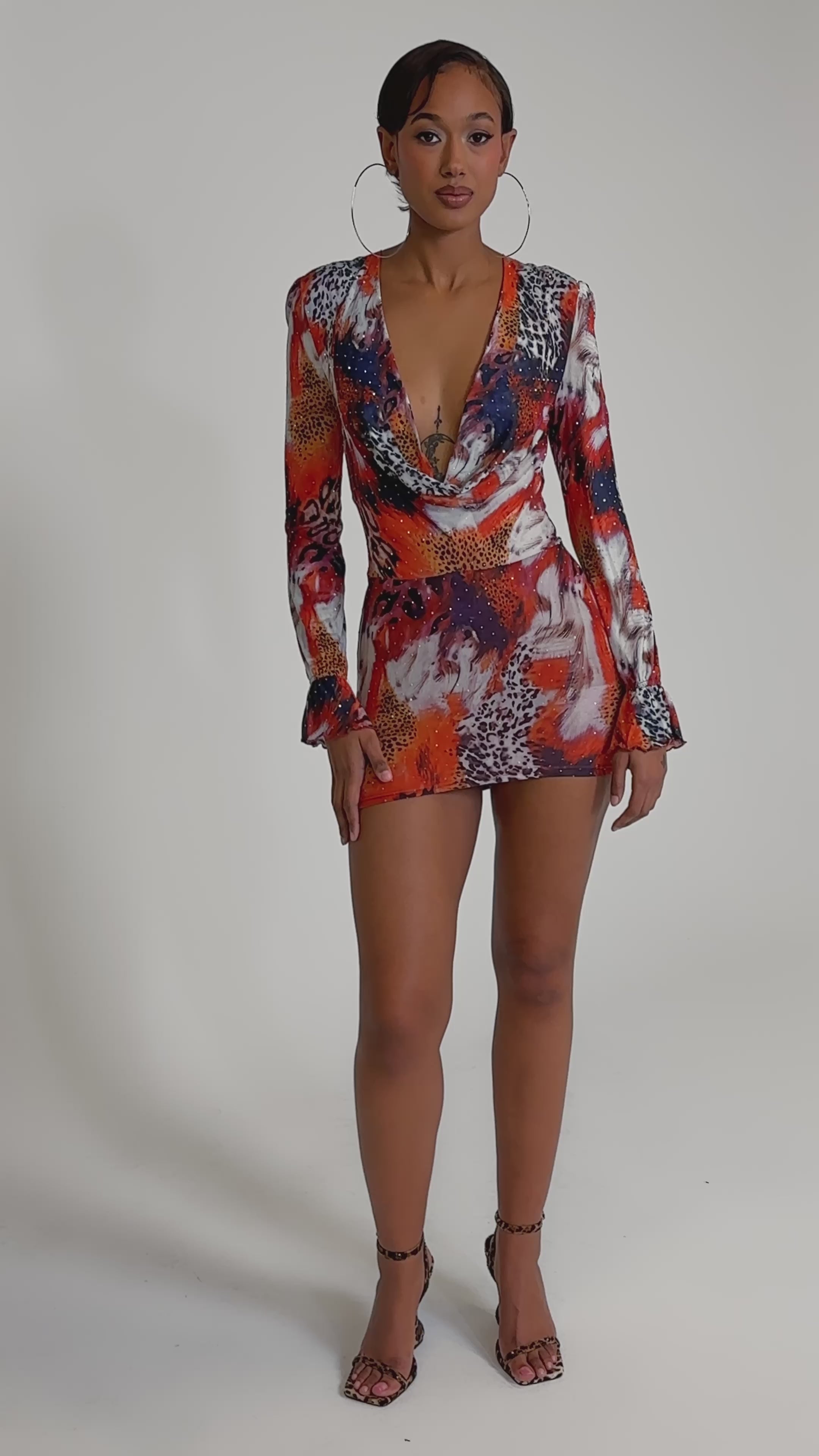 A video of a woman wearing the ON THE PROWL LEOPARD MINI DRESS with a bold red, yellow, and white leopard print. She pairs it with hoop earrings and strappy heels, her hair slicked back for a confident look.