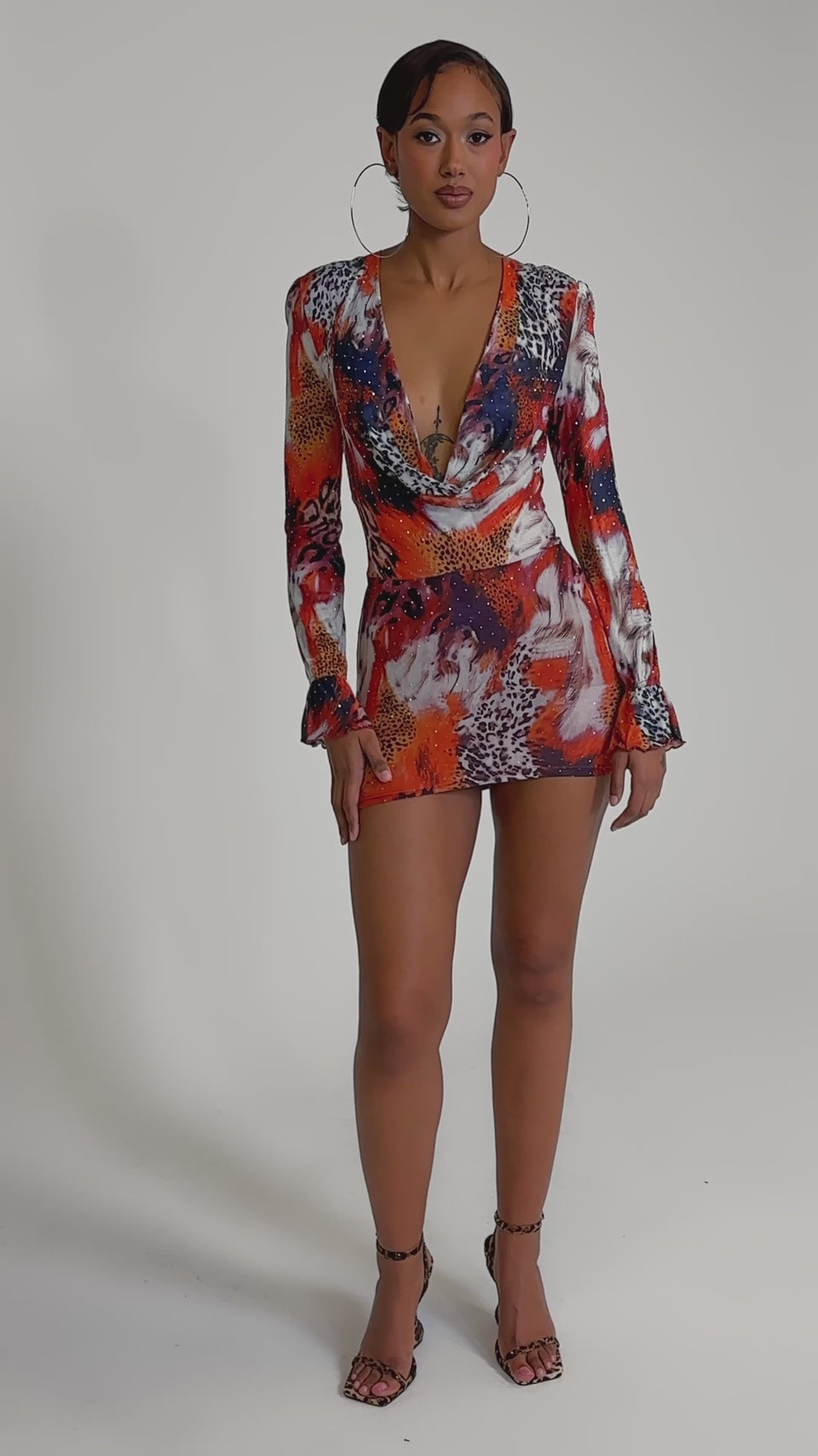 A video of a woman wearing the ON THE PROWL LEOPARD MINI DRESS with a bold red, yellow, and white leopard print. She pairs it with hoop earrings and strappy heels, her hair slicked back for a confident look.