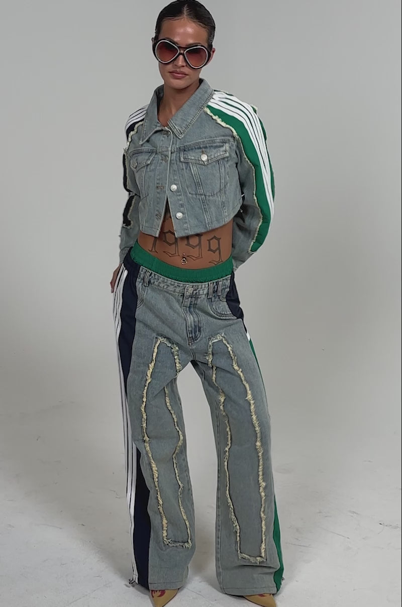 Video of woman modeling the ALL AROUND THE WORLD DENIM JACKET—an oversized cropped piece with windbreaker panel details and striped sleeve accents. She sports large sunglasses and a “1992” tattoo on her torso.