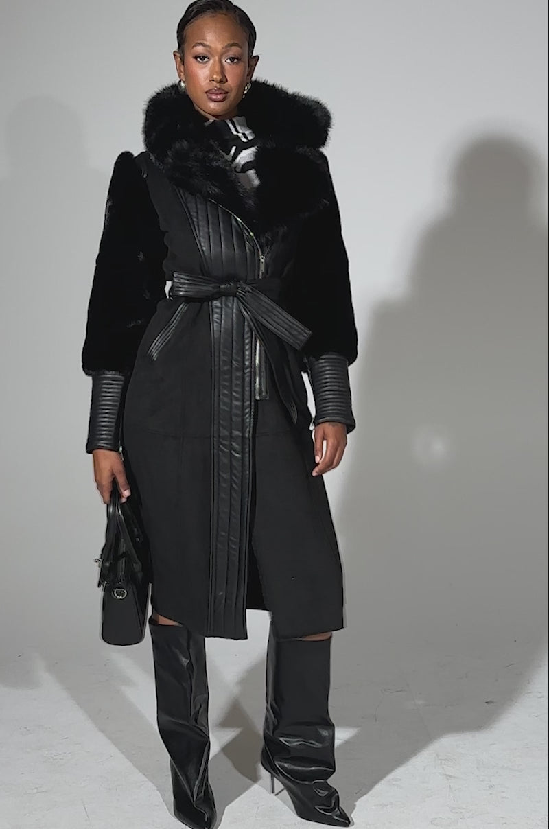 A video of a woman in the GOODIE MIXED MEDIA COAT, a black faux fur coat with a fur collar, paired with black knee-high boots and a small black handbag, her hair styled back against a plain light background.