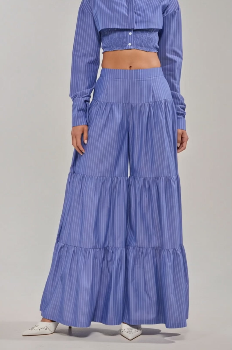 A video of a model wearing the BUSINESS ONLY STRIPE PALAZZO PANT, a high-rise, wide-leg blue striped design with three ruffled tiers, paired with white pointed shoes and a matching long-sleeved top for a statement look.