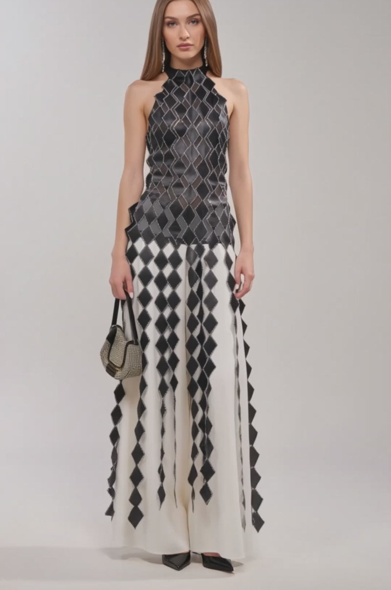 Video of woman modeling the ELINA SATIN TOP under a sleeveless, black and white geometric-patterned dress with a high neckline and layered diamond shapes, styled with a small handbag and black heels.