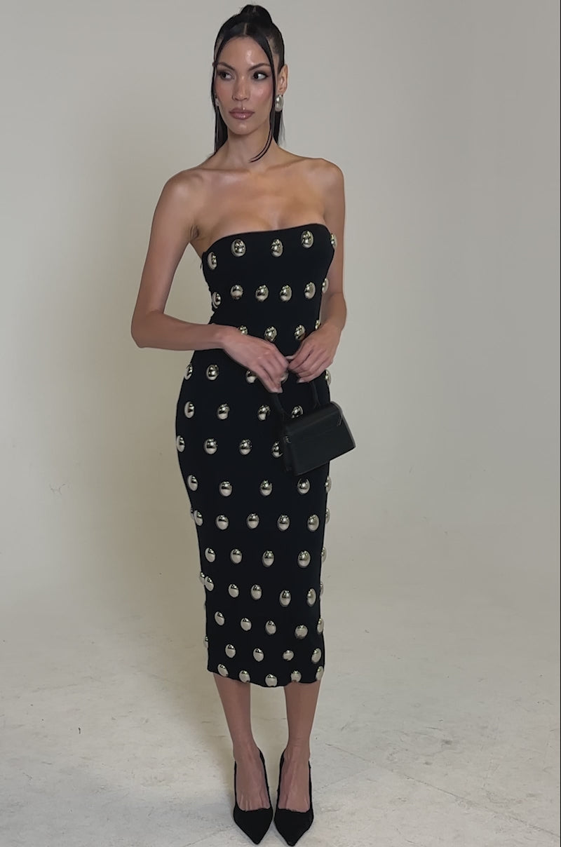 A video of a woman posing against a plain white background wearing the TIME AFTER TIME MIDI DRESS WITH METAL DETAILING, styled with a small black purse and black pointed heels.