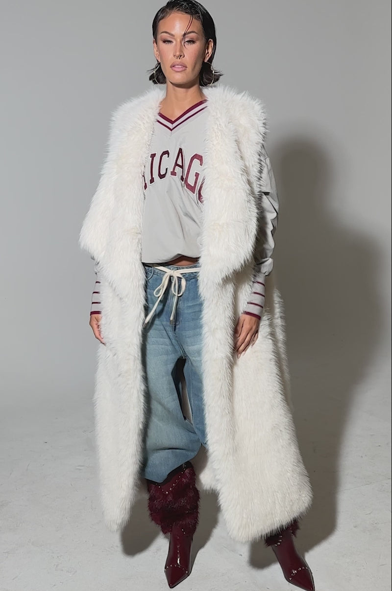 Video of woman modeling the DOWN TIME FAUX FUR VEST IN IVORY over a gray sweater with red text, distressed blue jeans, and burgundy boots.