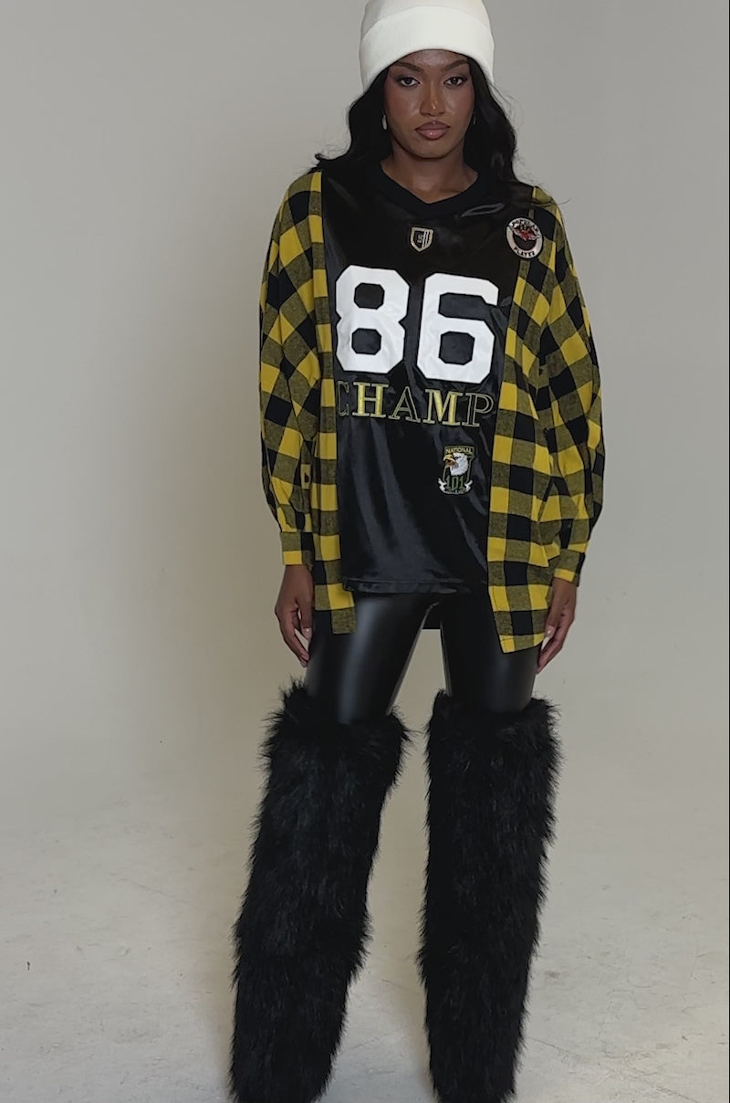 A video of a woman posing in a white beanie, streetwear oversized black and yellow checkered jacket, NEW CHAMP REBUILT JERSEY FLANNEL TOP, shiny black leggings, and tall black furry boots against a light gray backdrop.